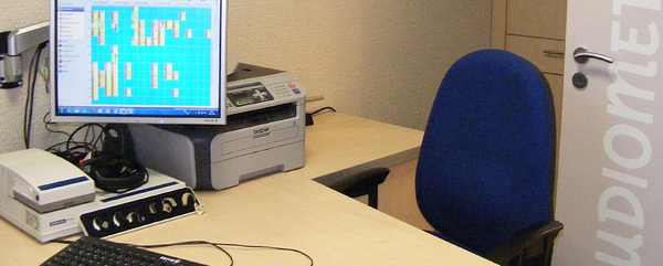 Modernes Hörtest-Arbeitsplatz mit Audiometer, Computer, Drucker und blauem Stuhl.